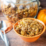 A bowl of pumpkin granola in front of a glass jar of it makes a cozy fall breakfast scene.