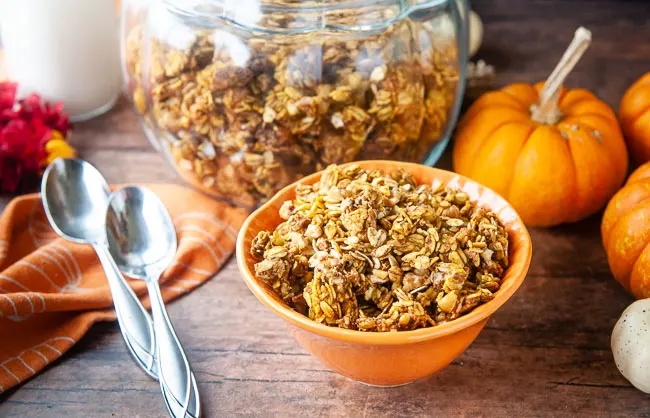 A bowl of pumpkin granola in front of a glass jar of it makes a cozy fall breakfast scene.