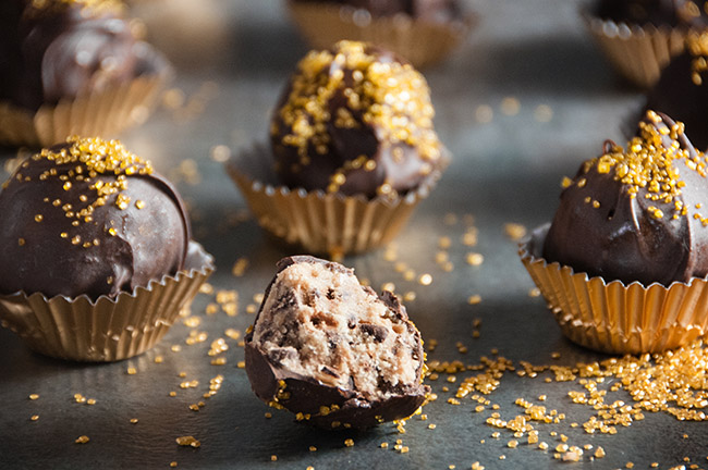 Chocolate Covered Cookie Dough Bites wrapped in gold wrappers with gold sprinkles. One with a bite out of it.