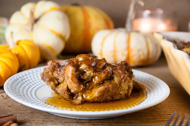 A slice of pumpkin French Toast Casserole on a white plate with syrup and pumpkins in the background