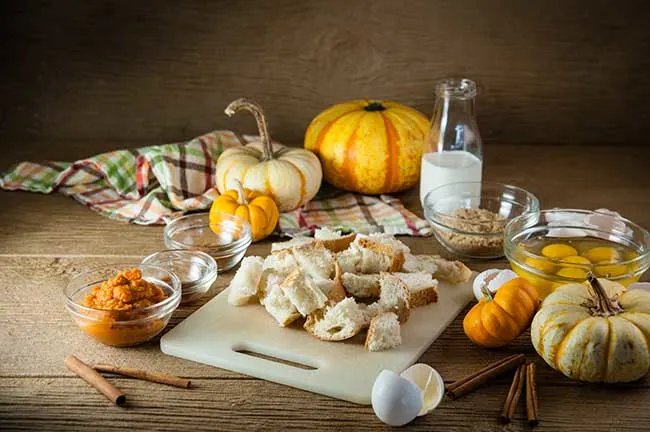 Ingredients for Pumpkin French Toast Casserole including bread cubes, cracked eggs, pumpkin puree, and spices laid out on a wooden table