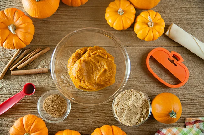 Pumpkin puree in a glass bowl surrounded by spices and brown sugar in glass bowls, with pumpkins and pie crust