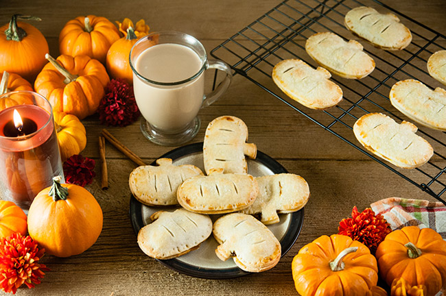 Pumpkin hand pies on a plate on a wood table with coffee, pumpkins, flowers, a candle, and cinnamon sticks