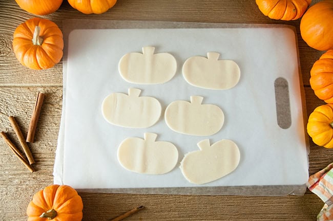 Pie crust in pumpkin shapes on a cutting board, surrounded by pumpkins and cinnamon sticks.