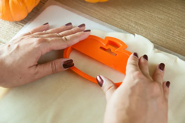 Woman's hands pressing an orange pumpkin cookie cutter into unrolled pie dough on a wood table