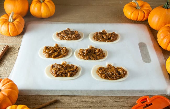 Pumpkin filling on pie crusts on a cutting board on a wood counter surrounded by small pumpkins