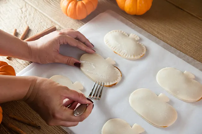 A woman's hands pressing a fork into the sides of a pumpkin shaped pies to seal the pies.