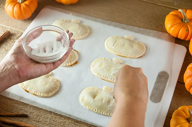 A woman's hands sprinkling sugar on pumpkin shaped pies.