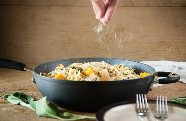 A hand sprinkling Parmesan cheese over a saute pan full of winter vegetable pasta