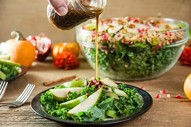 dressing being poured onto a plate of apple and pear kale salad sitting on a wood table with a large salad bowl and fall fruits behind it