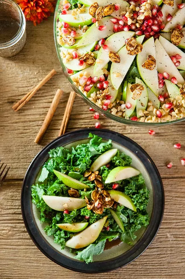 A plate of apple and pear kale salad sitting on a wood table with a large salad bowl and fall fruits behind it
