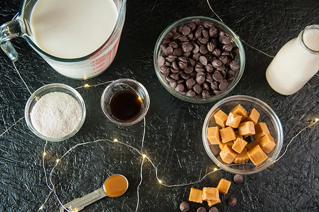 Milk, evaporated milk, sea salt, dark chocolate chips, caramels, vanilla all in bowls and containers on a black surface