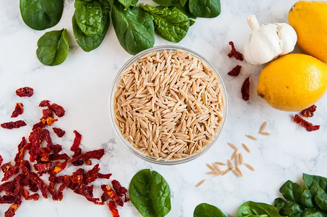 A bowl of orzo on a light background surrounded by lemons, garlic, spinach and sun dried tomatoes