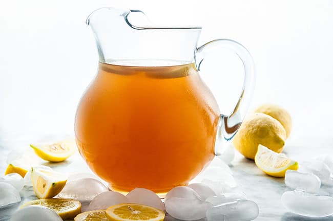 Homemade Iced Tea in a glass and glass pitcher with lemons and ice cubes on white