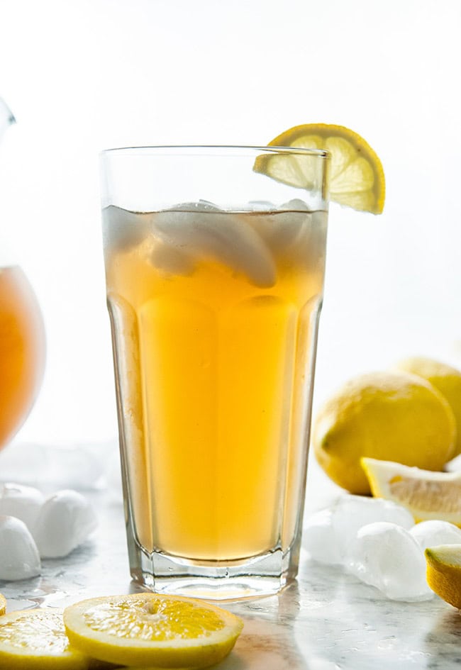 Homemade Iced Tea in a glass and glass pitcher with lemons and ice cubes on white