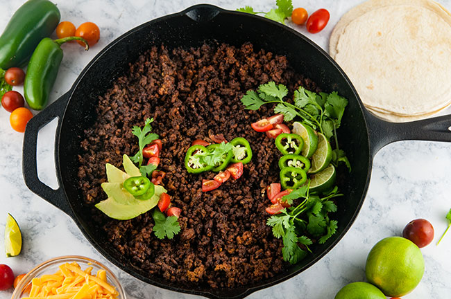 Skillet of taco meat garnished with jalapenos, avocado, tomatoes, and cilantro on light background