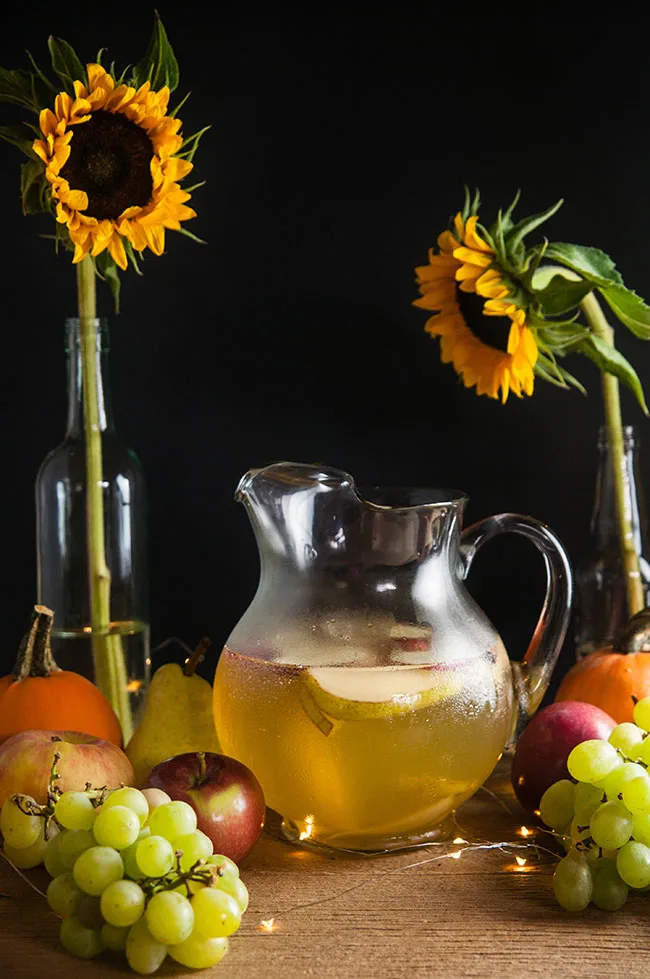 A pitcher of sangria with apples and pears on a wood table surrounded by harvest fruits, sunflowers, and twinkle lights