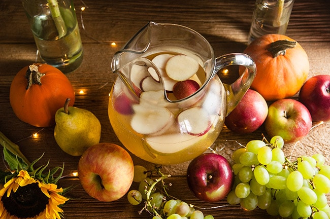 A pitcher of sangria with apples and pears on a wood table surrounded by harvest fruits, sunflowers, and twinkle lights