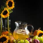 A pitcher of sangria with apples and pears on a wood table surrounded by harvest fruits, sunflowers, and twinkle lights