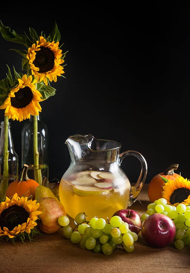 A pitcher of sangria with apples and pears on a wood table surrounded by harvest fruits, sunflowers, and twinkle lights