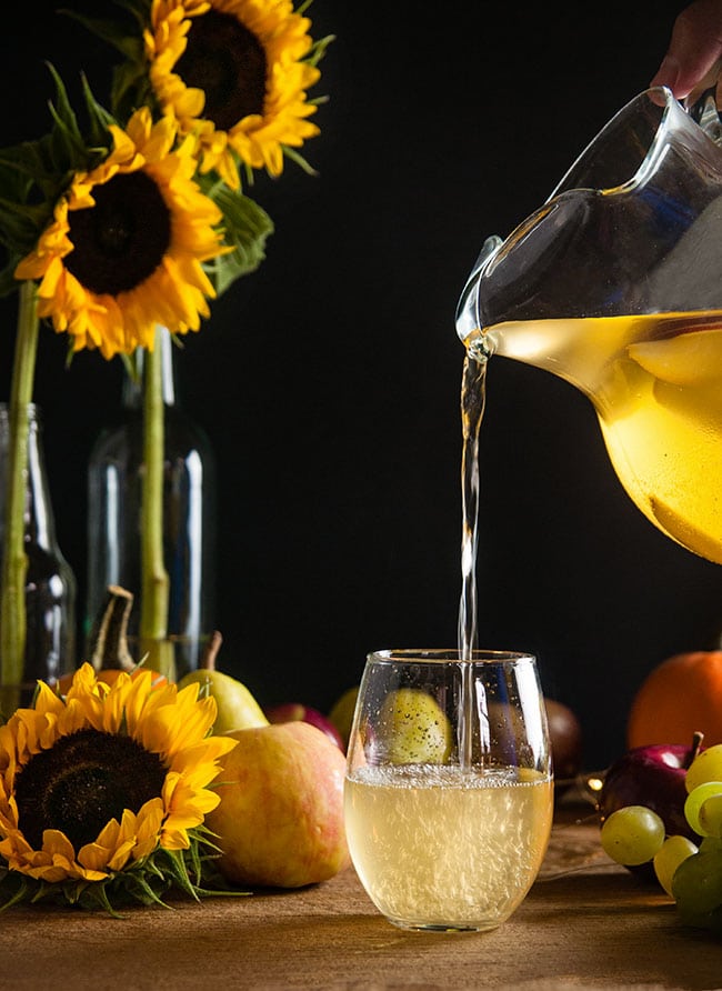 White apple sangria in a pitcher being poured into a stemless wineglass on wood table with sunflower and harvest fruits