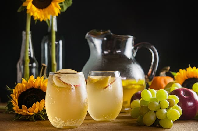 A pitcher of sangria with apples and pears on a wood table surrounded by harvest fruits, sunflowers, and twinkle lights