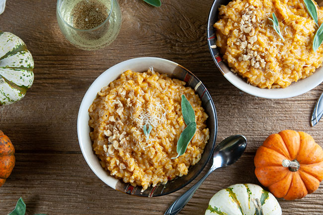 2 bowls of pumpkin risotto on wood with small pumpkins, glasses of white wine, and parmesan cheese