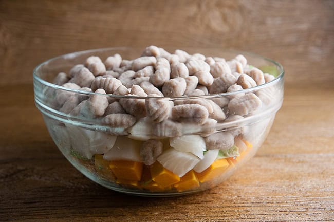 a bowl of gnocchi with chopped veggies on wood