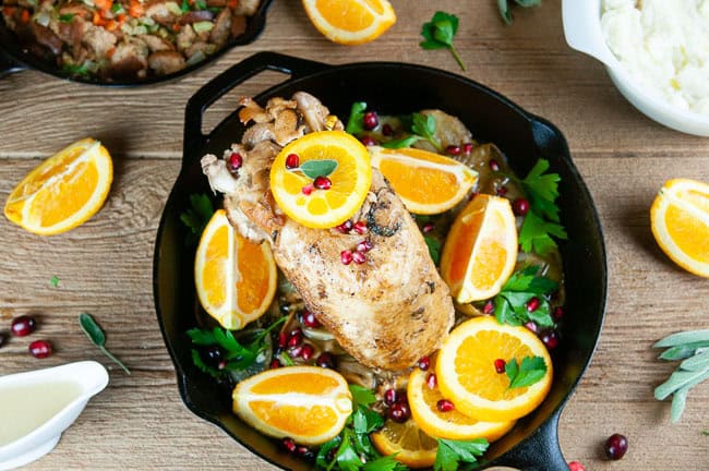 Garlic roasted turkey breast in a cast iron skillet with orange slices and greens on wood surrounded by dishes of mashed potatoes, stuffing, and gravy