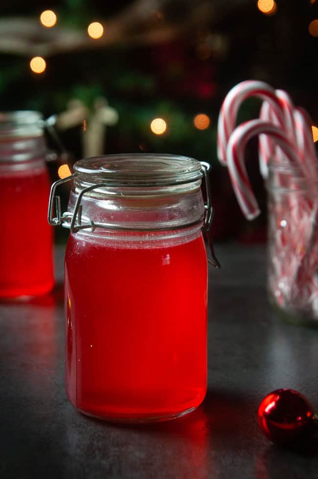 2 jars of red candy cane vodka on dark in front of Christmas trees and candy canes