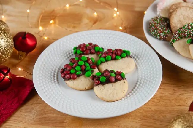 A plate of sugar cookies dipped in chocolate and M&Ms on wood with holiday ornaments and lights store bought sugar cookie dough hacks