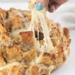 A woman's hand pulling a square of bread from a loaf of French onion pull apart bread. Cheese pull.