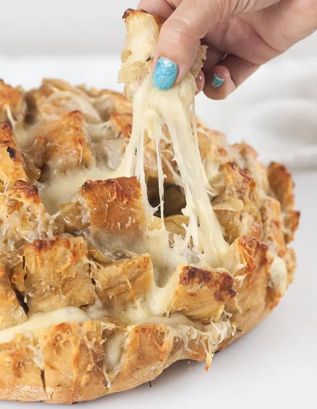 A woman's hand pulling a square of bread from a loaf of French onion pull apart bread. Cheese pull.