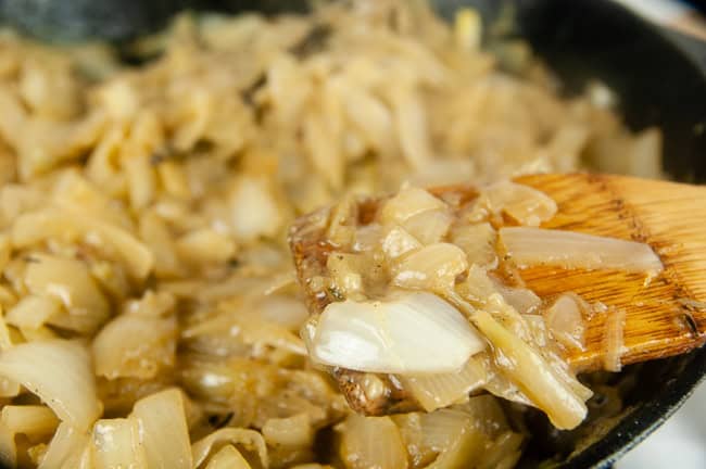 A white softened onion slice on a spatula in front of a skillet of caramelized onions