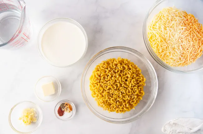 Ingredients for Instant Pot Mac and Cheese are macaroni, heavy cream, water, butter, spices, garlic, cheese and hot sauce. They are shown in glass bowls in a white kitchen.