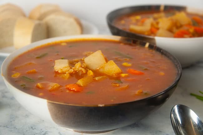 two bowls of Instant Pot Vegetable Soup with Brown Rice on white marble with a plate of sliced baguette and spoons