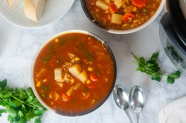 two bowls of Instant Pot Vegetable Soup with Brown Rice on white marble with a plate of sliced baguette and spoons