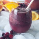 two pomegranate cranberry smoothies on white marble with ice cubes, orange slices, and frozen cranberries