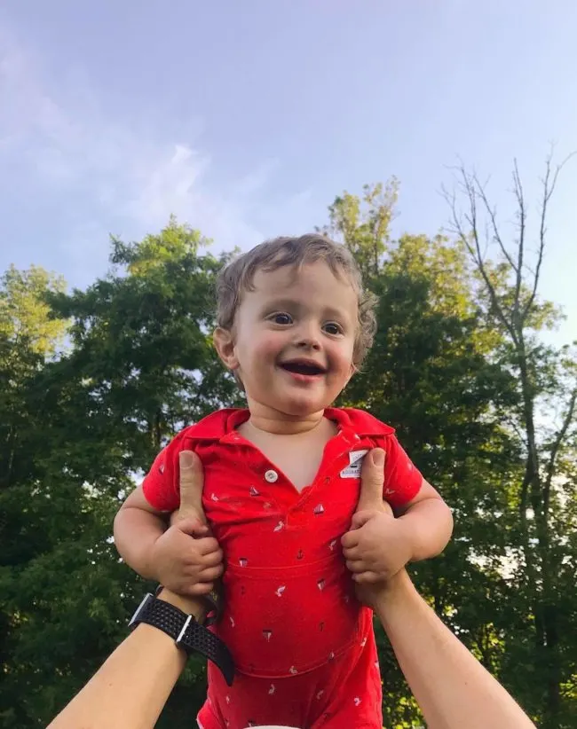 Cute baby being held up in front of trees and smiling