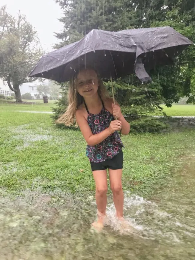 Little girl wearing a bathing suit and holding a black umbrella standing in shin deep water running through a yard as it pours