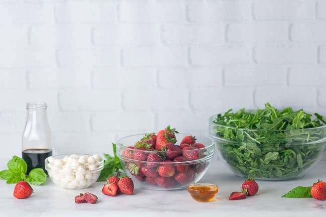 Ingredients for Strawberry Caprese Salad: arugula, strawberries, mini mozzarella balls, basil, balsamic vinegar, and honey