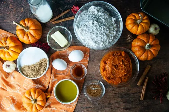 Small bowls of ingredients for Cinnamon Swirl Pumpkin Bread with Streusel Topping on a wood table: flour, sugar, pumpkin, eggs, oil, butter, spices, and milk