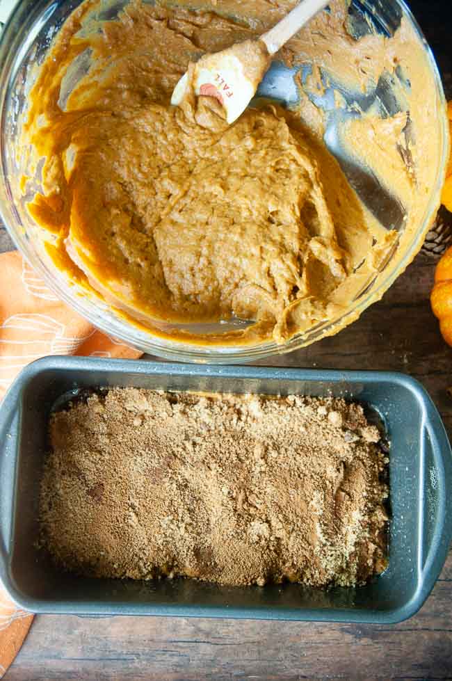 The pumpkin bread is shown with the cinnamon sugar layer sitting on a half filled loaf pan with a clear bowl full of the remaining batter next to it.