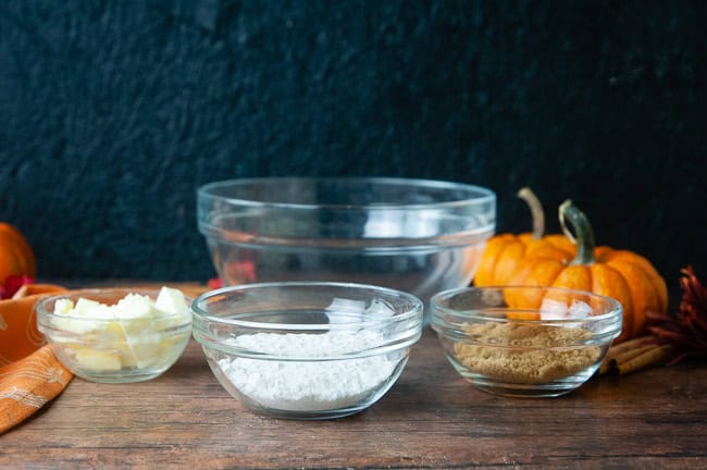 The ingredients for the streusel topping (flour, cold cubes of butter, sugar, spices) are arranged in bowls on a wood table with pumpkins.