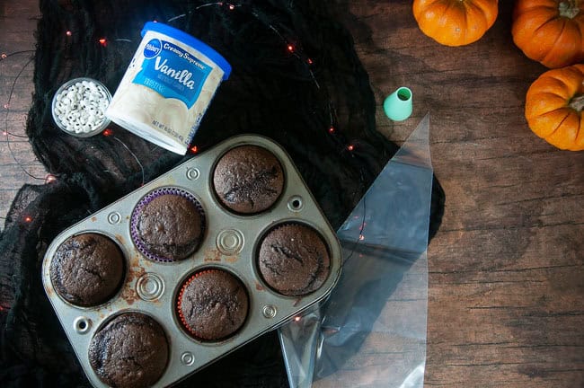 The ingredients for ghost cupcakes are chocolate cupcakes, vanilla frosting, candy eyes, and black decorator gel. They are show with a piping bag and large round icing tip.