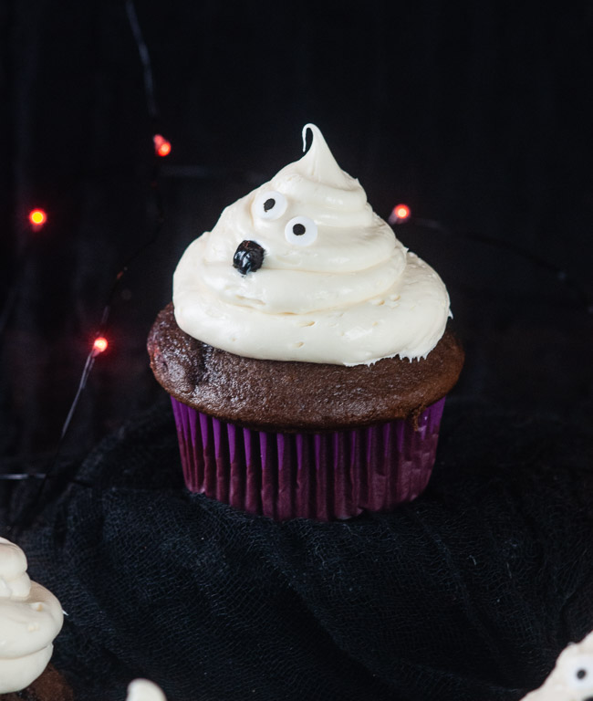 Ghost cupcakes on a black background with orange twinkle lights make a bootiful treat for Halloween.