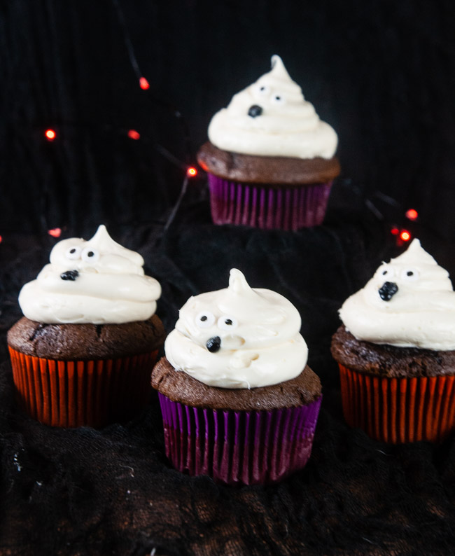 Ghost cupcakes on a black background with orange twinkle lights make a bootiful treat for Halloween.
