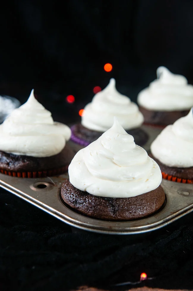 A large dollop of vanilla icing makes the ghosts on the cupcakes. Vanilla iced cupcakes in a cupcake tin on black.