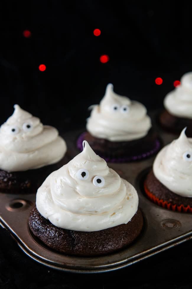 Decorate the ghosts with candy eyes and black decorator gel mouths. Ghost cupcakes in a cupcake tin on black with orange twinkle lights.