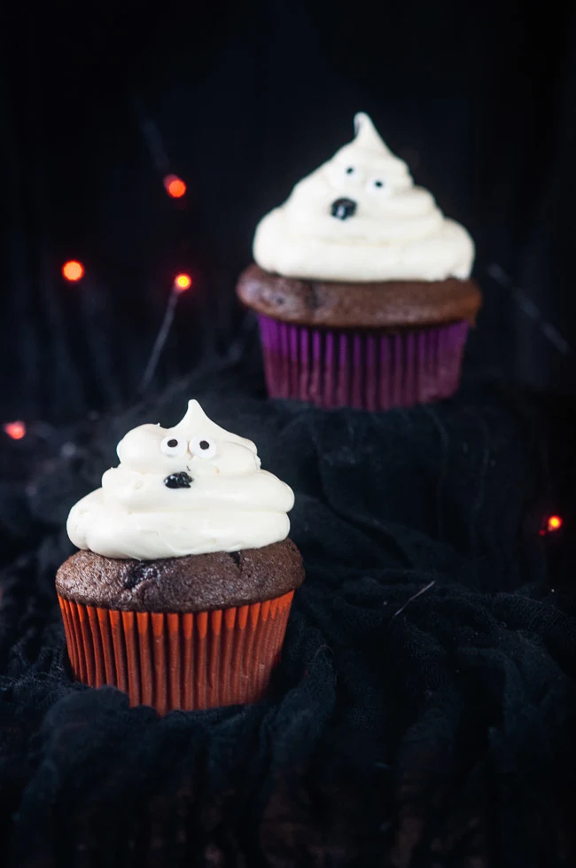 Two ghost cupcakes on a black background with orange twinkle lights make a bootiful treat for Halloween.
